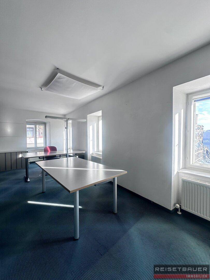 Helles Büro mit Teppichboden, mehreren Arbeitsplätzen und großen Fenstern.