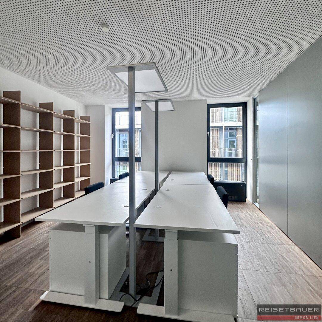 Bürobereich mit zwei Reihen von Schreibtischen, großen Fenstern und einem hohen Bücherregal.