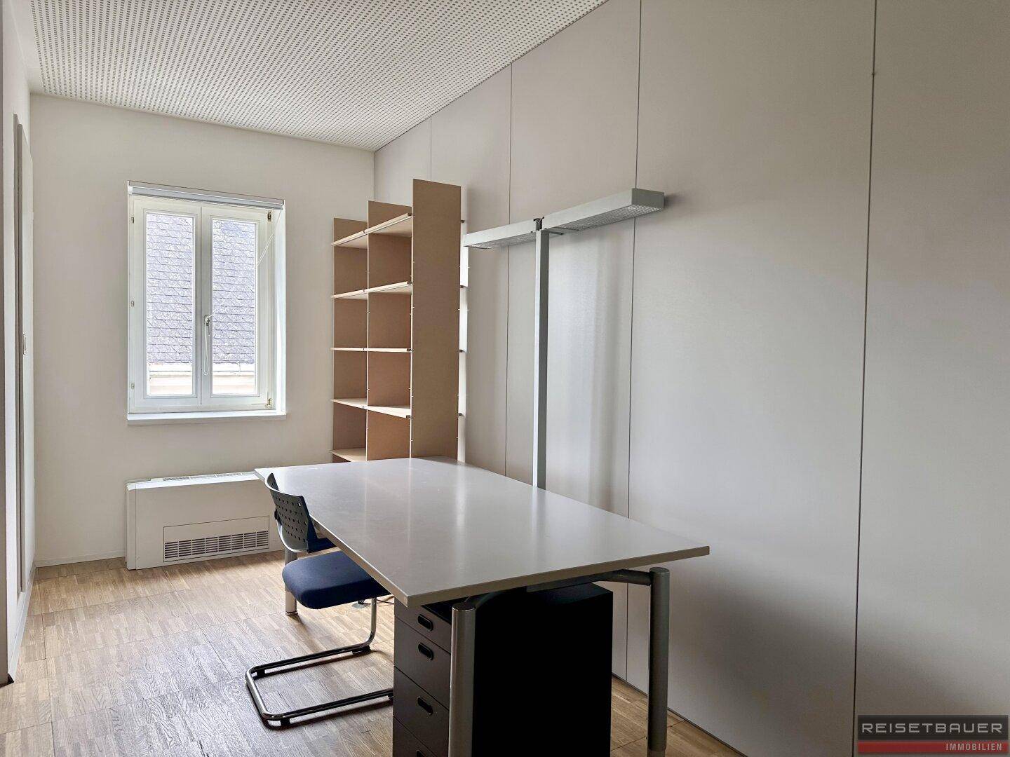 Einzelbüro mit Holzboden, Regal, Schreibtisch und einem Fenster mit Blick nach draußen.