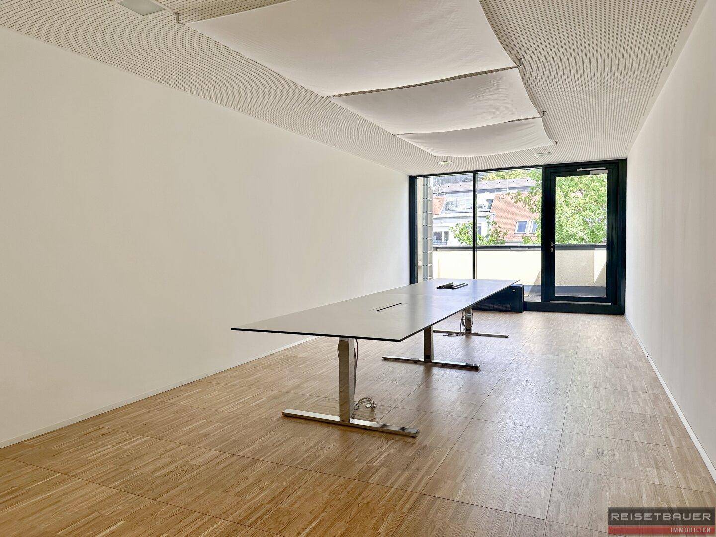 Großer Konferenzraum mit Holzboden, langen Tischen und bodentiefen Fenstern mit Zugang nach draußen.
