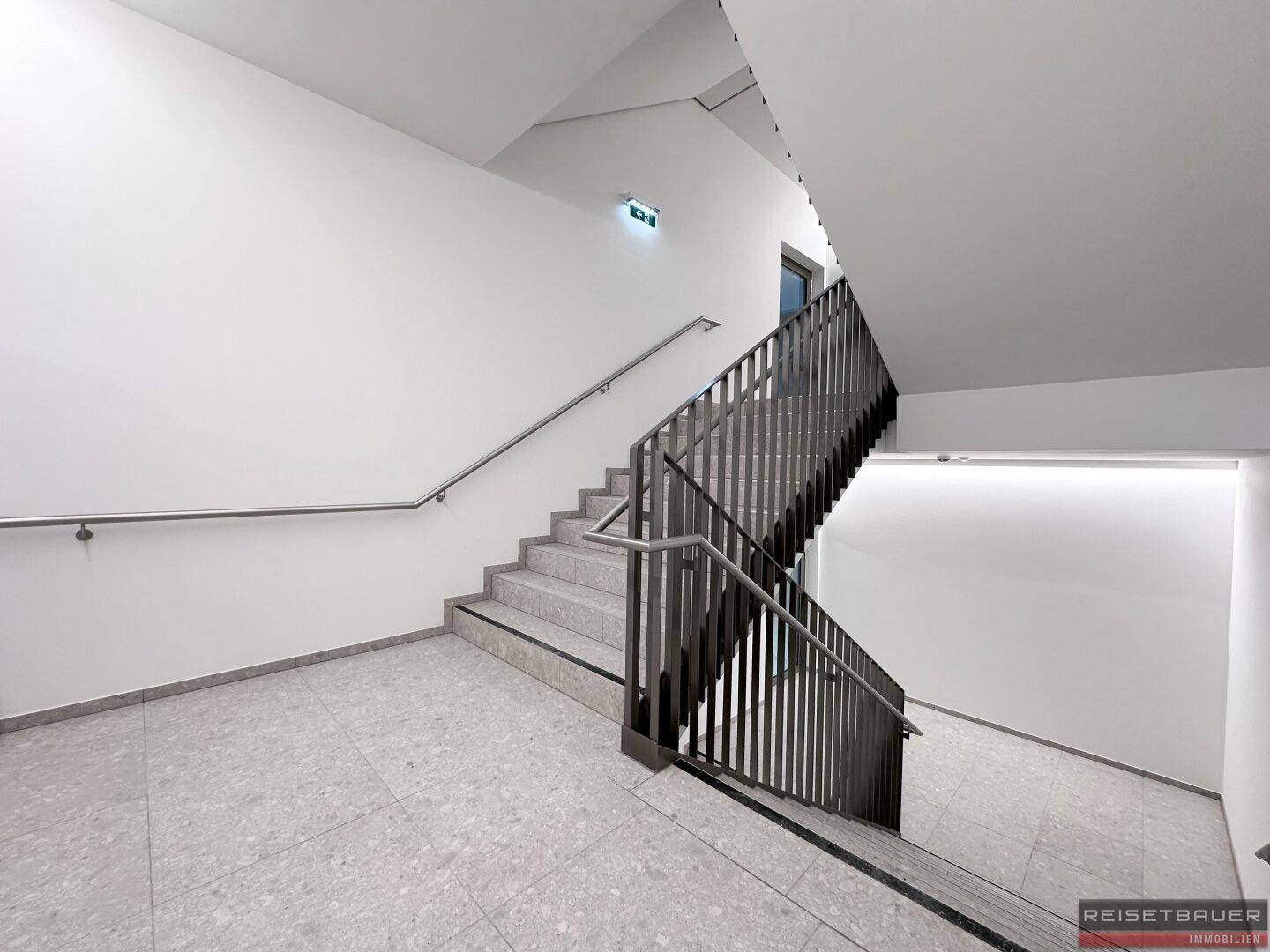 Modernes Treppenhaus mit Metallgeländer, hellen Wänden und Fliesenboden, gut beleuchtet.