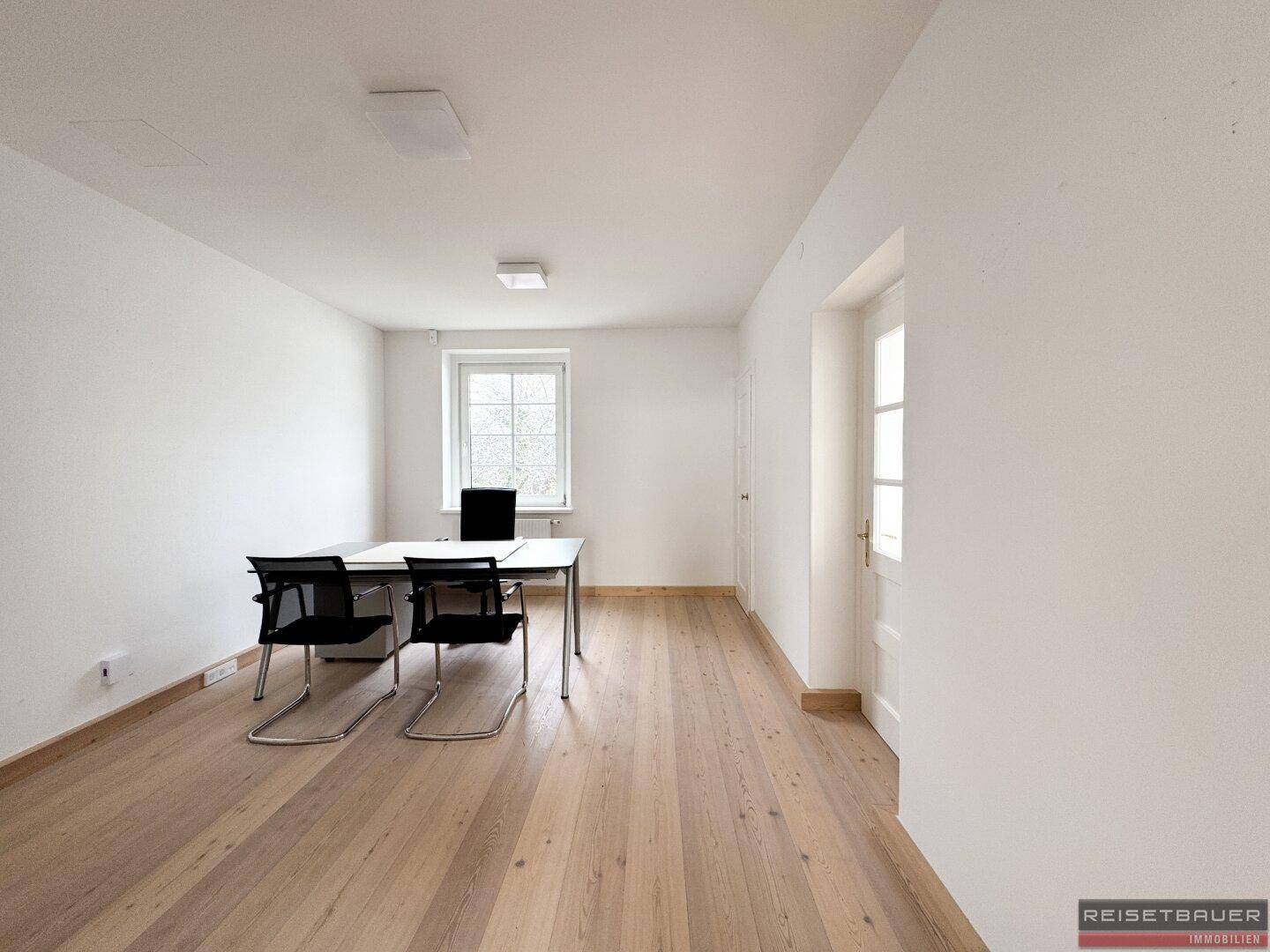 Kleiner Büroraum mit Schreibtisch und Stühlen, hellem Holzboden und Fensterblick.