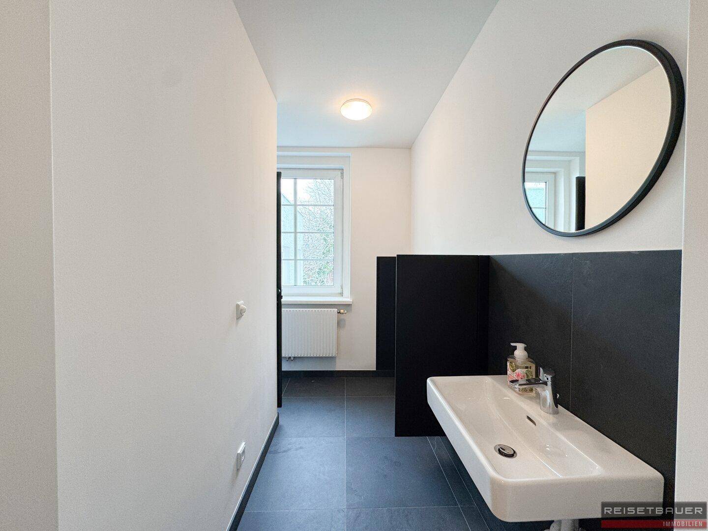 Modernes Badezimmer mit dunklen Fliesen, weißem Waschbecken und großem Spiegel.