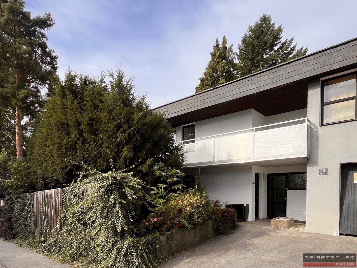Modernes Haus mit weißer Fassade, Balkon und umgebender Bepflanzung.