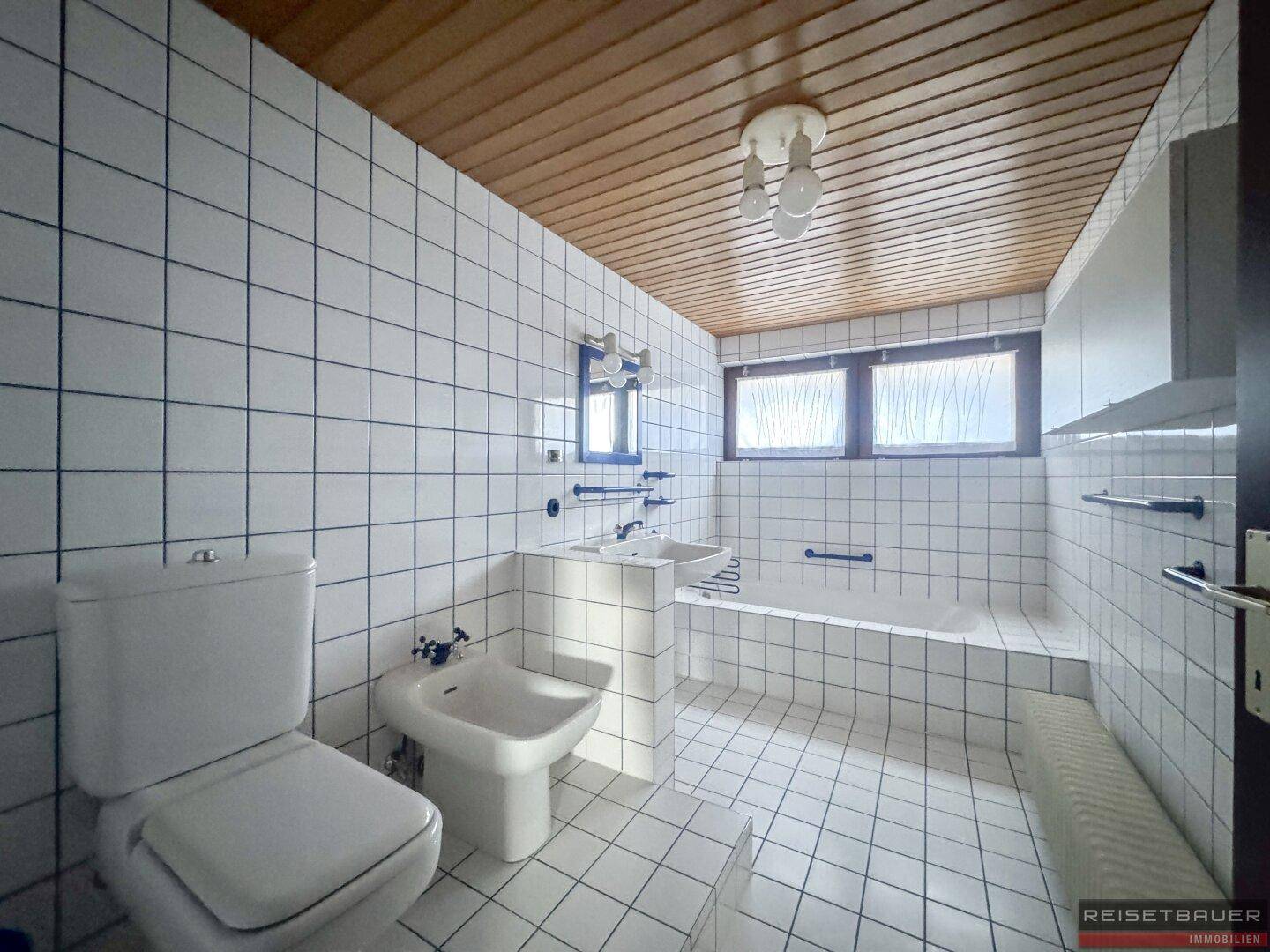 Badezimmer mit weißen Fliesen, Badewanne, Bidet und Holzdecke, funktional eingerichtet.