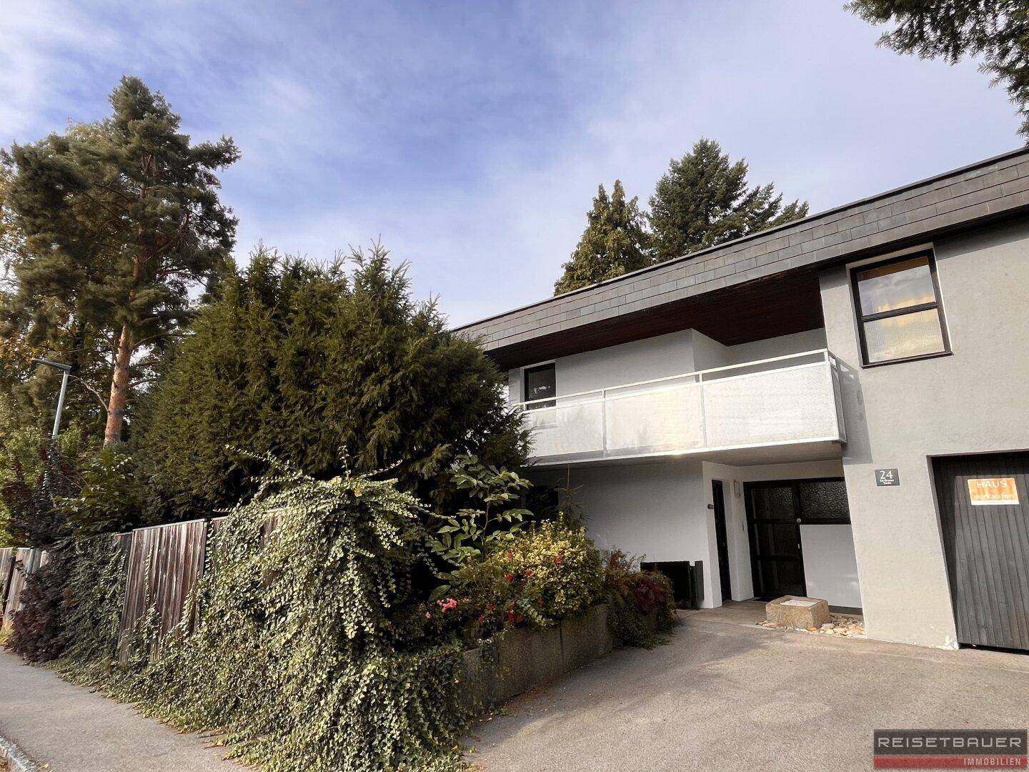Modernes Gebäude mit Balkon und Garage, umgeben von Bäumen und Sträuchern.