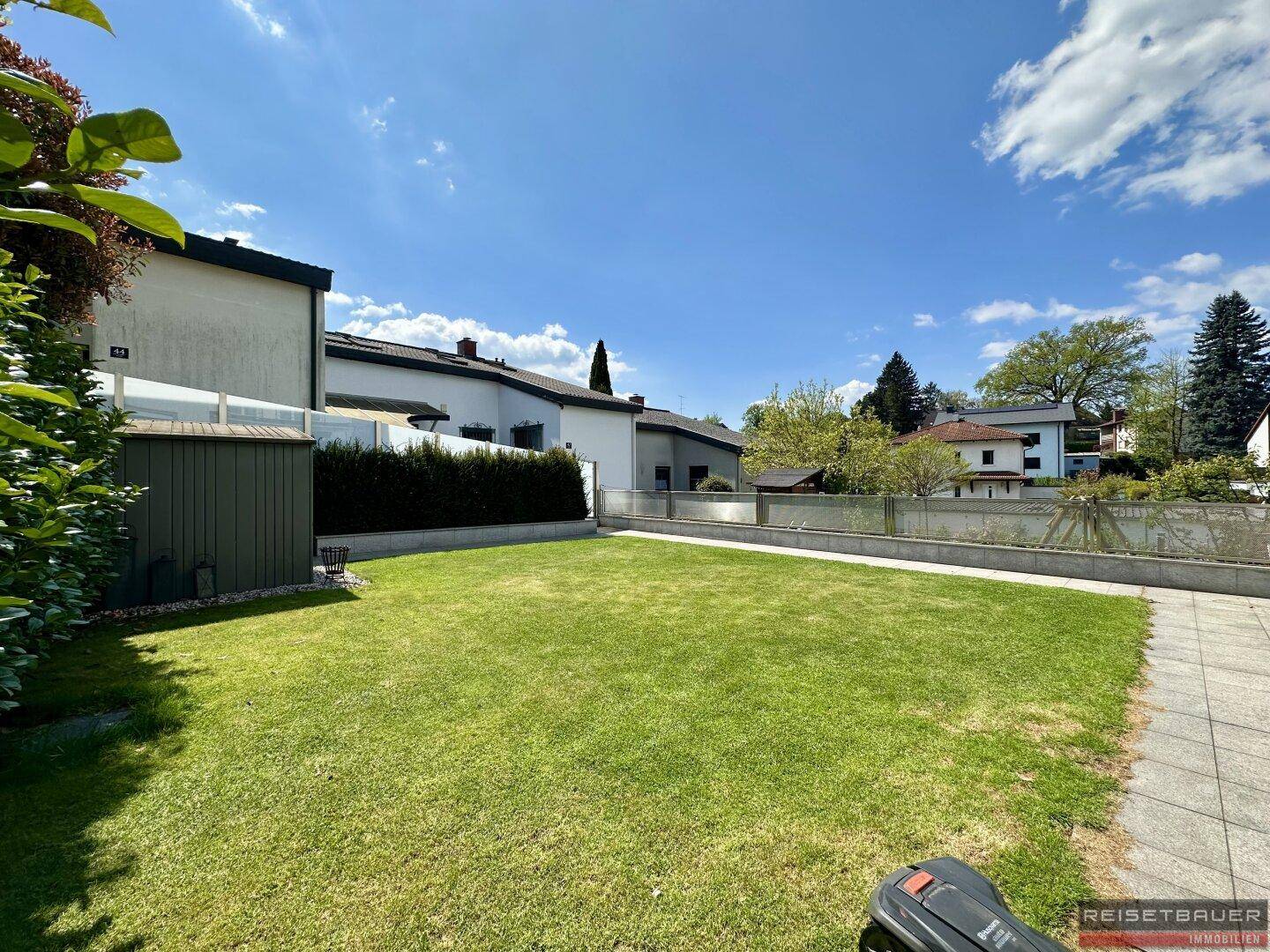 Weitläufiger Rasenbereich im Garten mit einem Mähroboter und Blick auf die umliegenden Häuser.