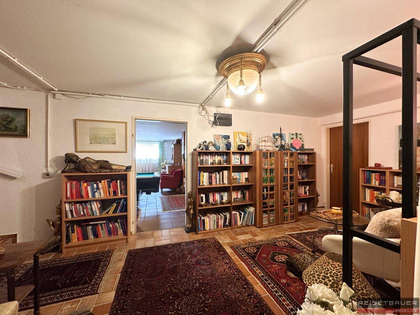 Heller Bibliotheksbereich mit mehreren Bücherregalen, Teppichen und Blick in einen angrenzenden Raum.