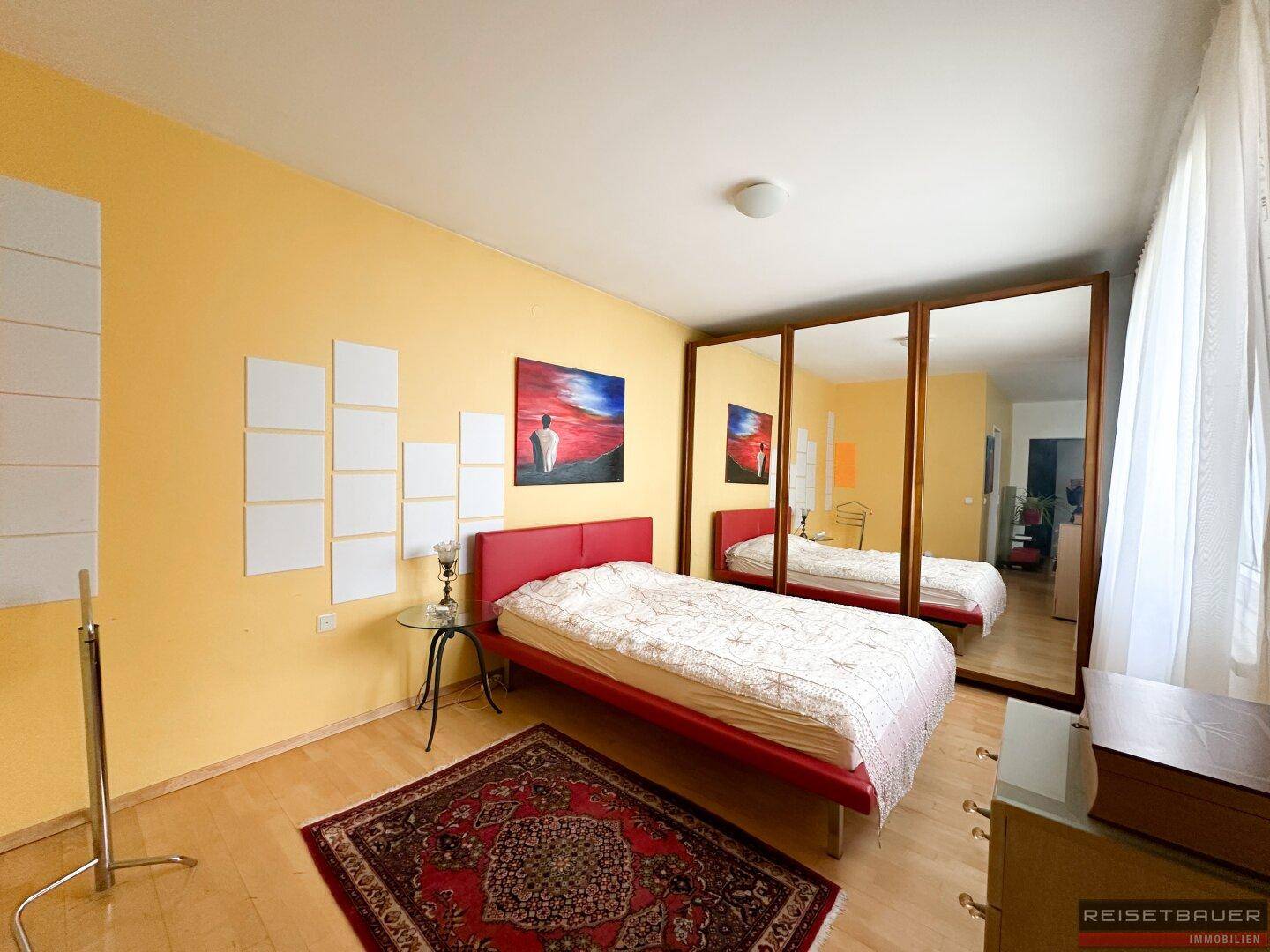 Helles Schlafzimmer mit gelben Wänden, rotem Bett und einem großen Spiegelschrank.