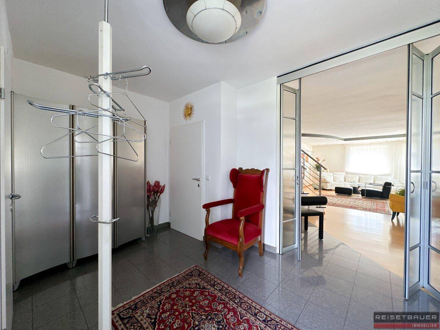 Heller Eingangsbereich mit Garderobe, rotem Sessel und Zugang zum Wohnbereich durch eine Glastür.