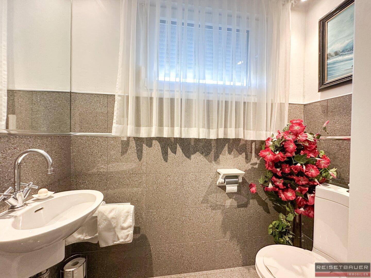 Kleines Badezimmer mit WC, Waschbecken, Fenster und grauen Fliesen an den Wänden.