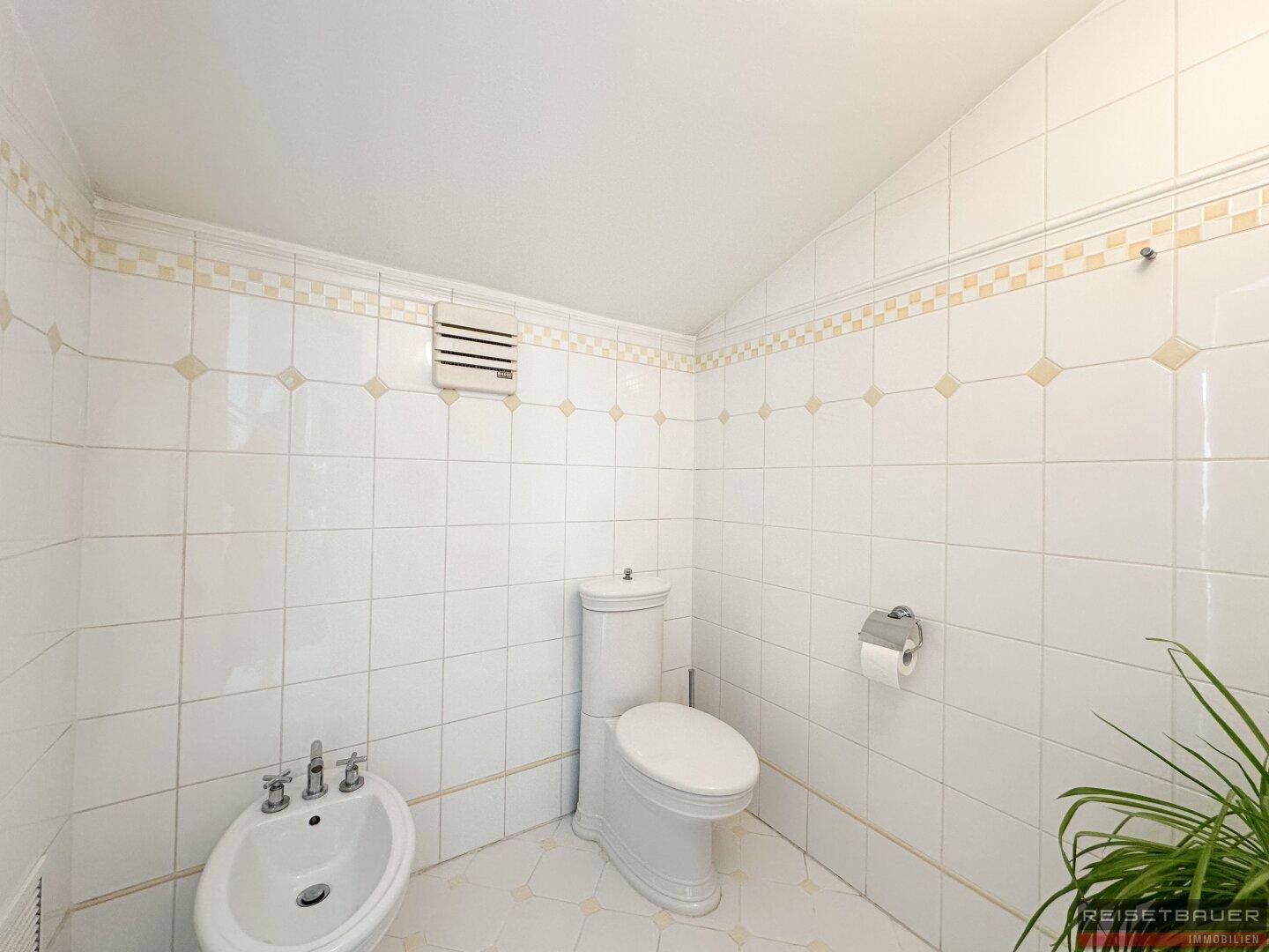 Helles WC mit weißen Fliesen, Toilette und Bidet, ausgestattet mit einem Lüftungssystem.