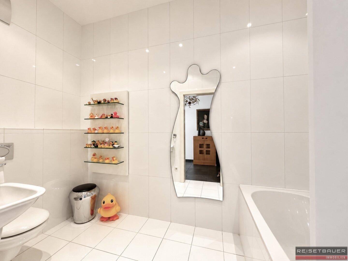 Weiß gefliestes Badezimmer mit Badewanne, WC und dekorativem Spiegel.