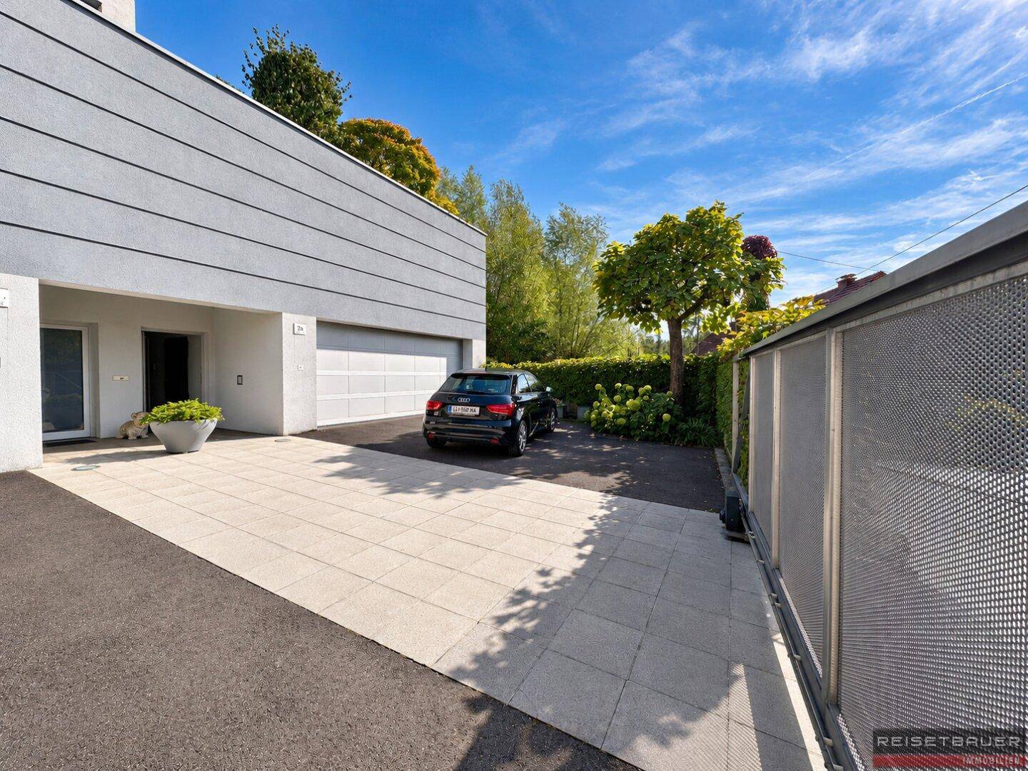 Moderne Gebäudefassade mit integrierter Garage und gepflasterter Zufahrt, umgeben von Grünflächen.