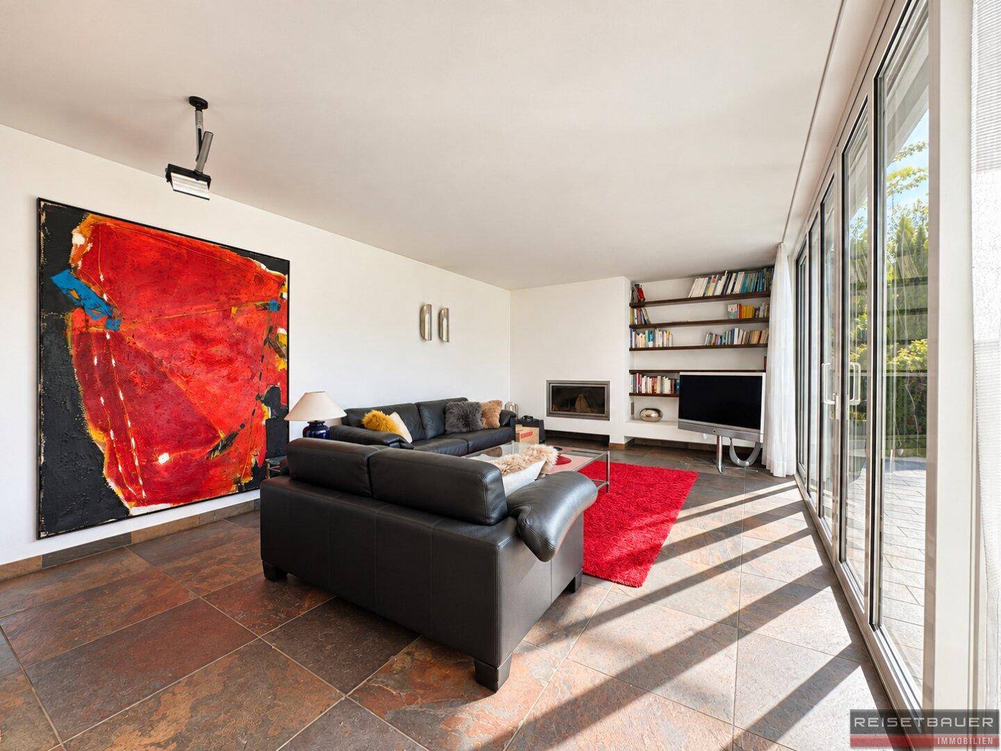 Großzügiges Wohnzimmer mit Kunstwerken, Kamin und viel Tageslicht durch die Fensterfront.