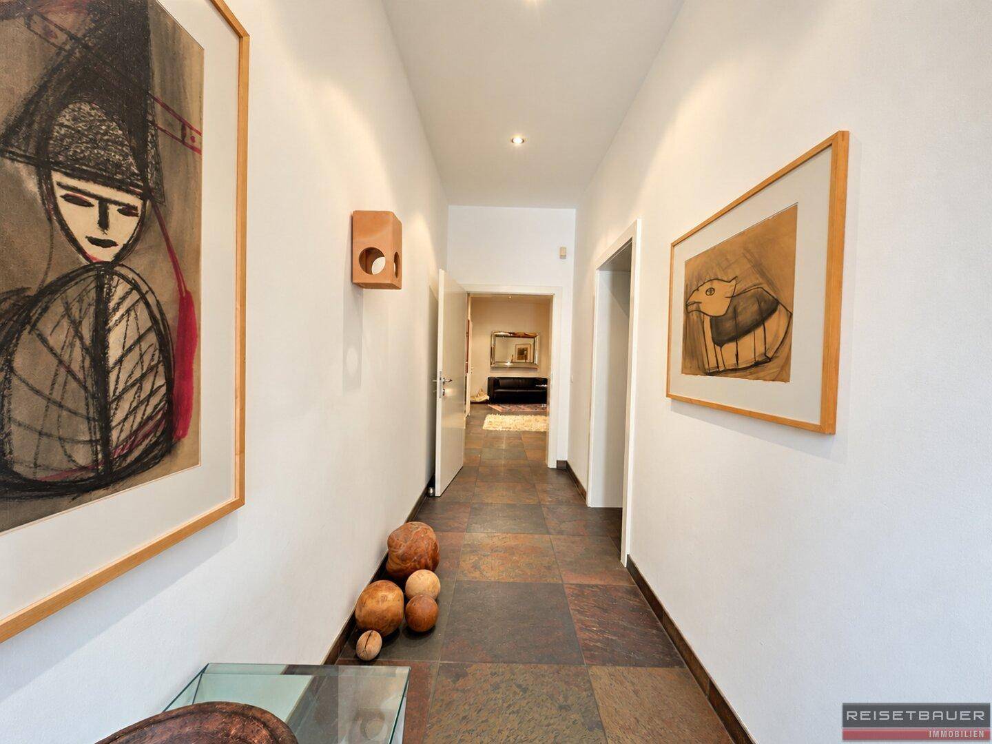 Langer Flur mit Natursteinfliesen, Kunstwerken und indirekter Beleuchtung.