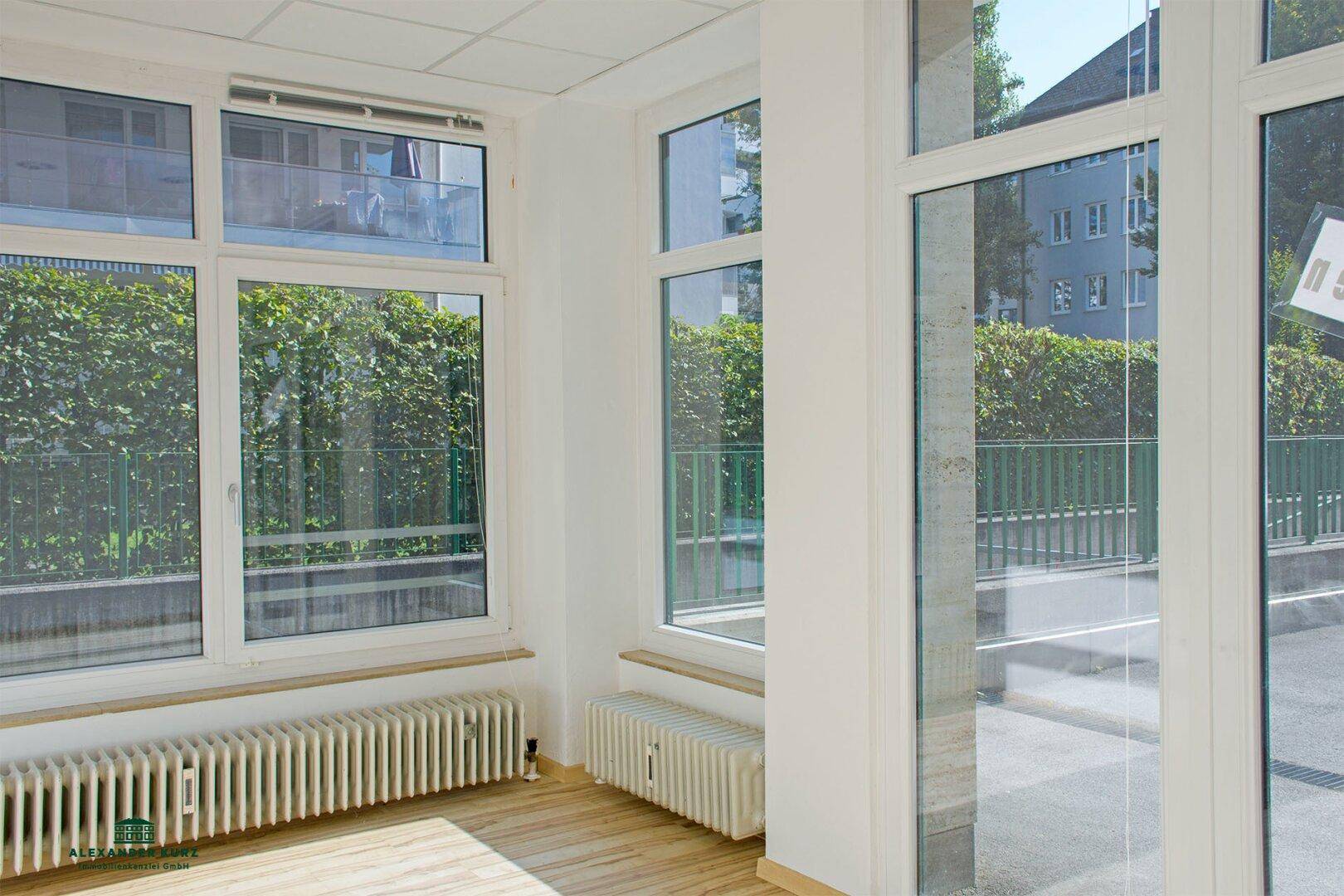 Heller Raum mit großen Fenstern, die auf eine grüne Hecke und die Straße blicken, mit Holzboden.