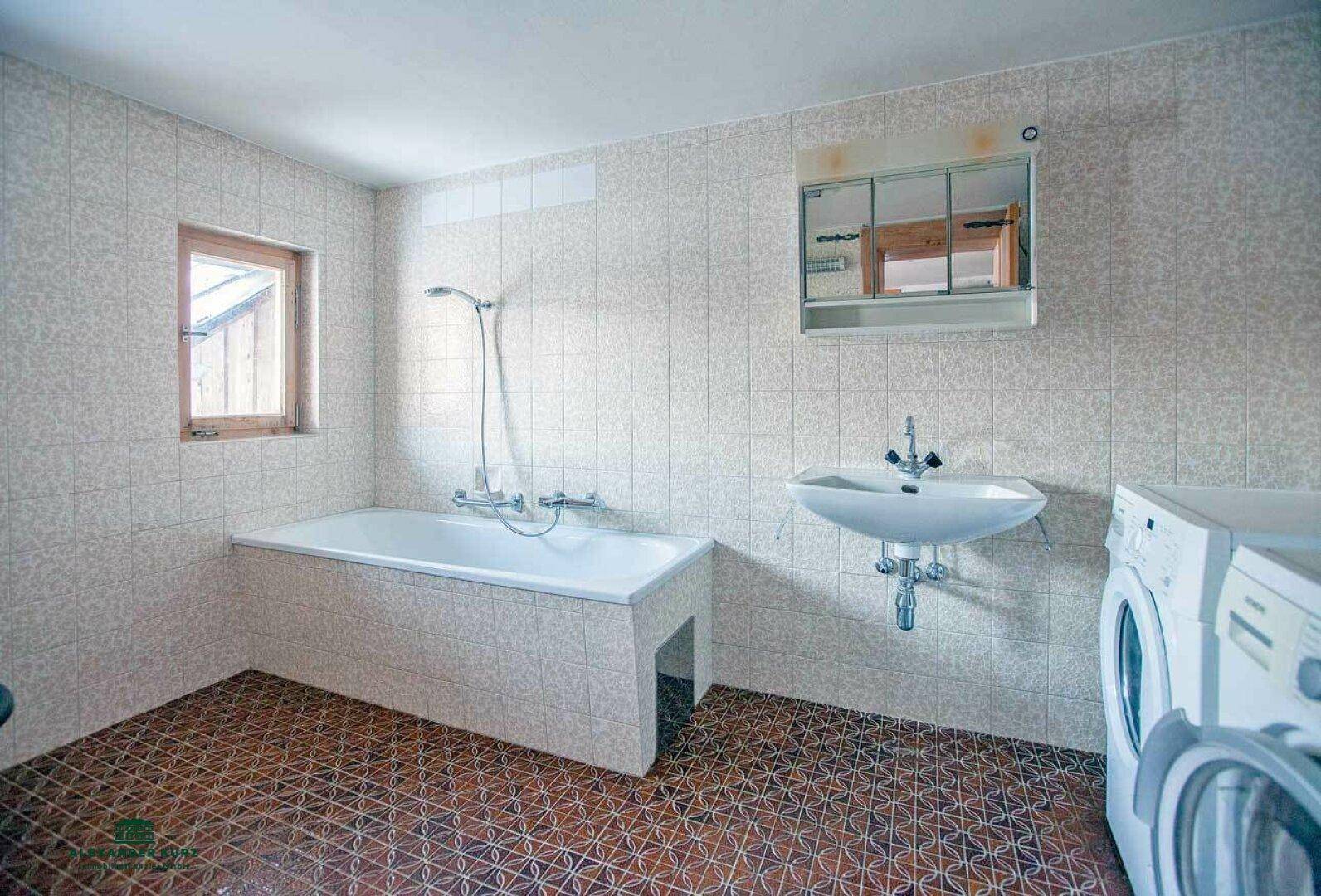 Gefliestes Badezimmer mit Badewanne, Waschbecken, Spiegelschrank und Waschmaschine.