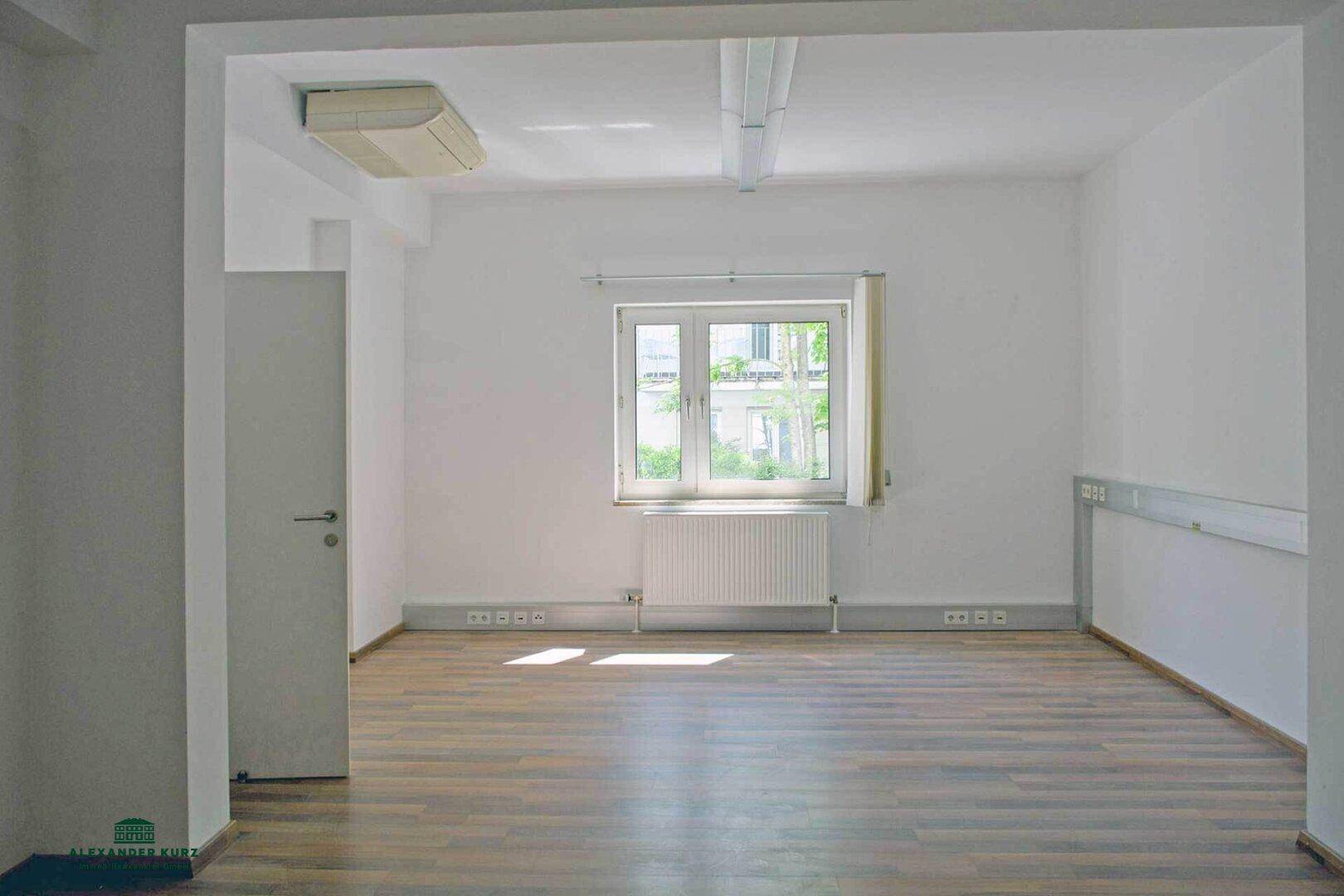Großer, leerer Raum mit Laminatboden, weißer Decke und Wänden, sowie einem Fenster für natürliche Belichtung.