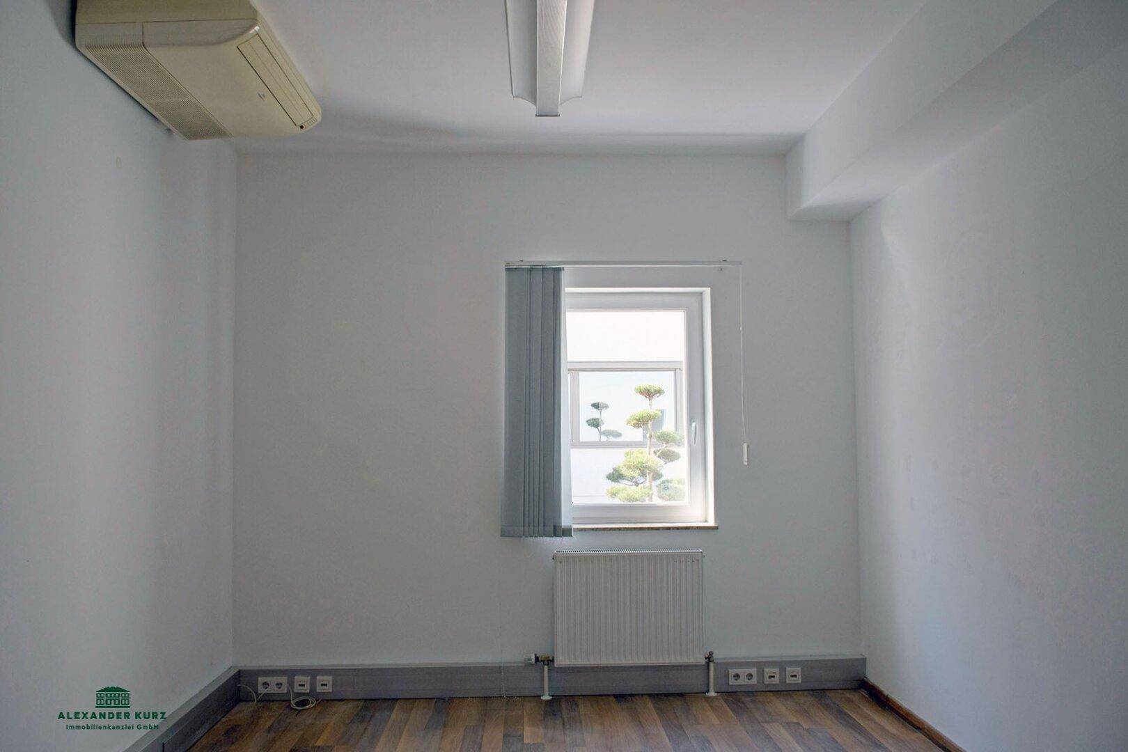 Leerer Raum mit Holzboden, Fenster, Heizkörper und einer Klimaanlage an der Decke.