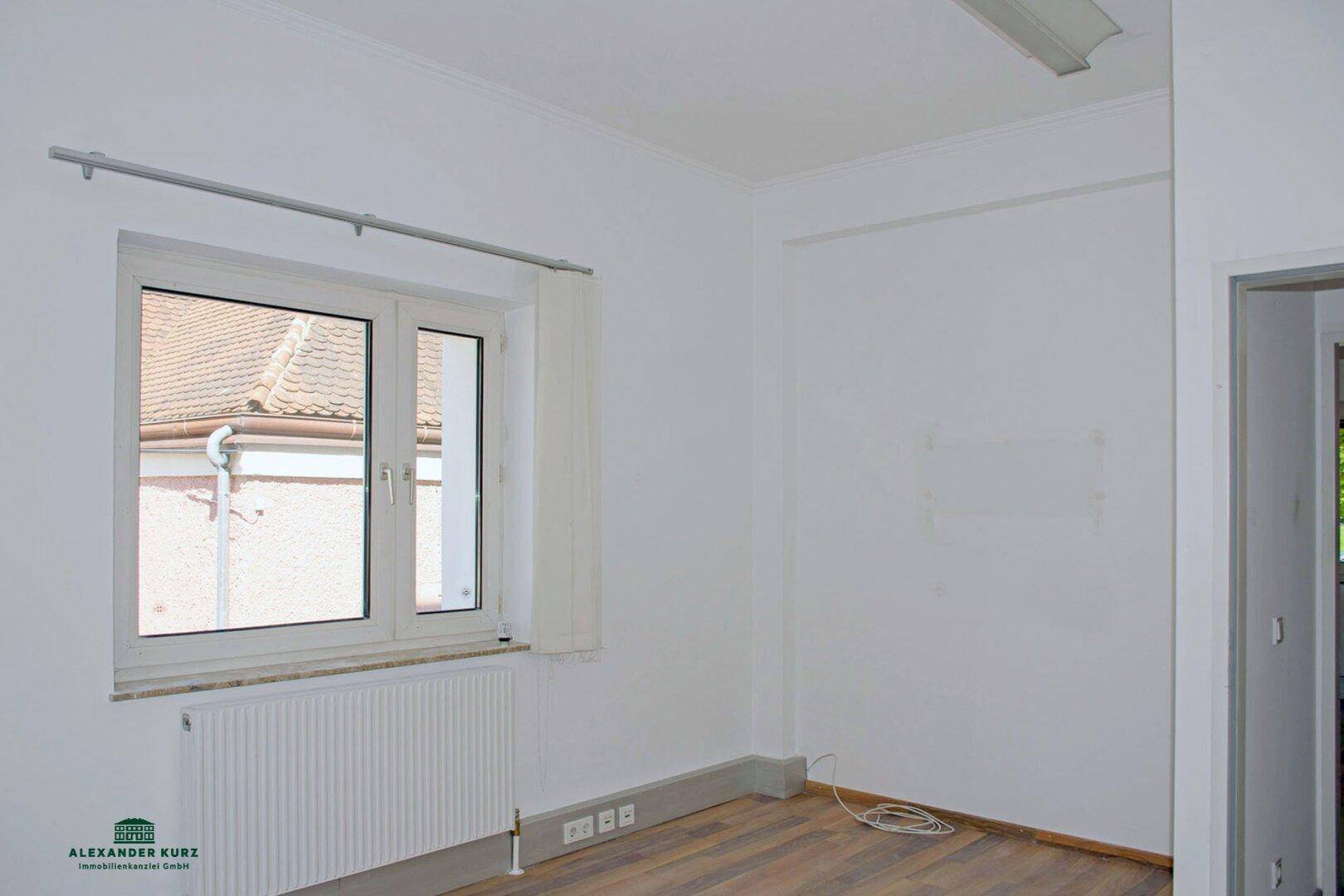 Leerer Raum mit Holzboden und einem Fenster, das den Blick auf ein Ziegeldach freigibt.