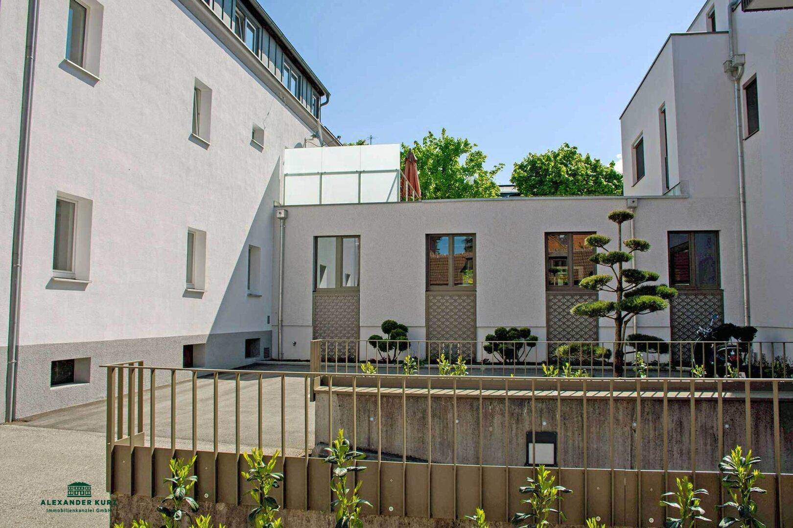 Außenansicht eines Gebäudekomplexes mit modernen Fassaden, kleinen Bäumen und einem Zaun.
