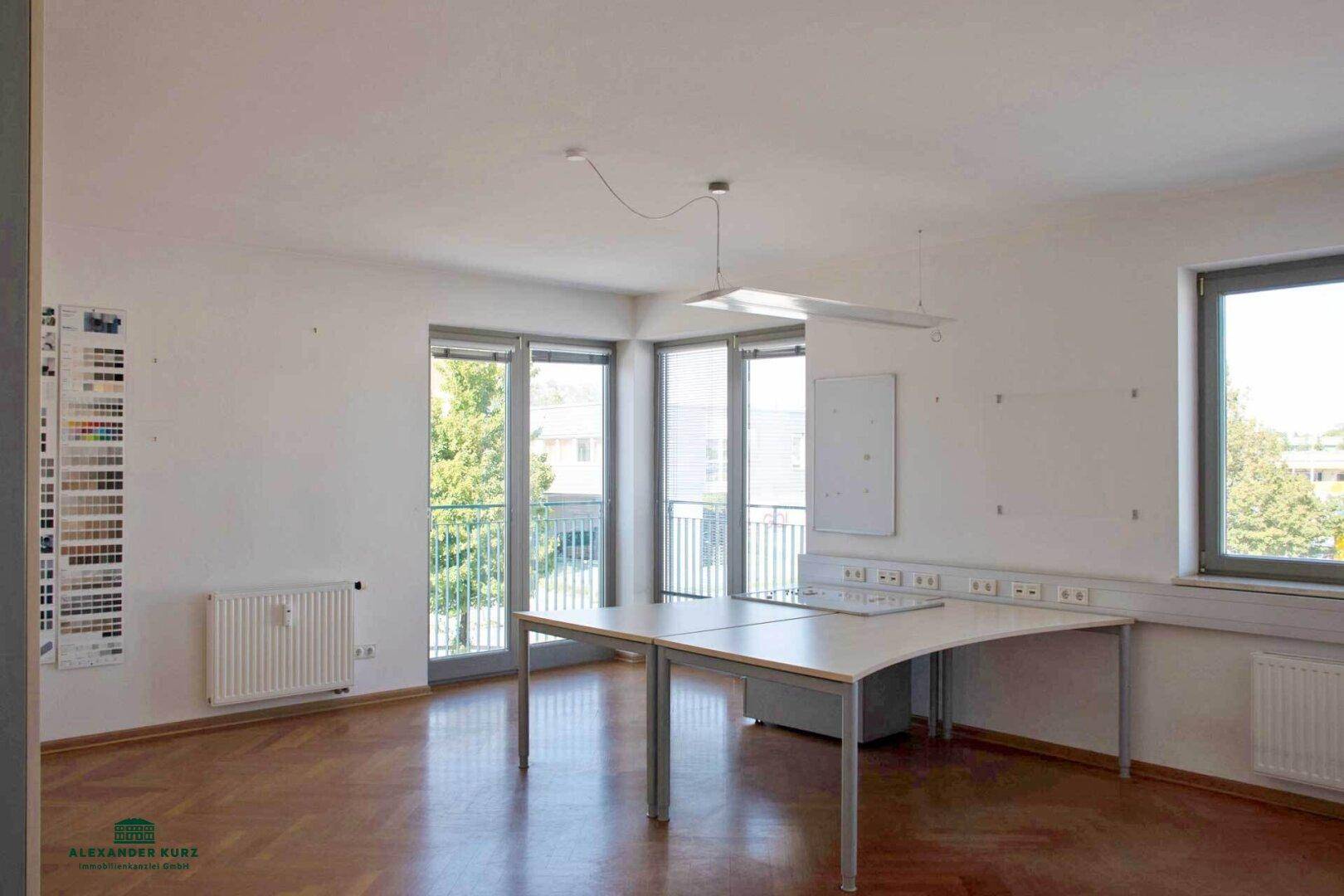 Helles Büro mit Parkettboden, großen Fenstern und Zugang zu einem Balkon mit Blick ins Grüne.