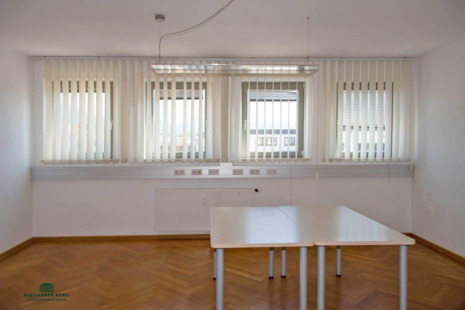 Büro mit mehreren Fenstern, vertikalen Jalousien und einem langen Arbeitstisch, ideal für Teamarbeit.