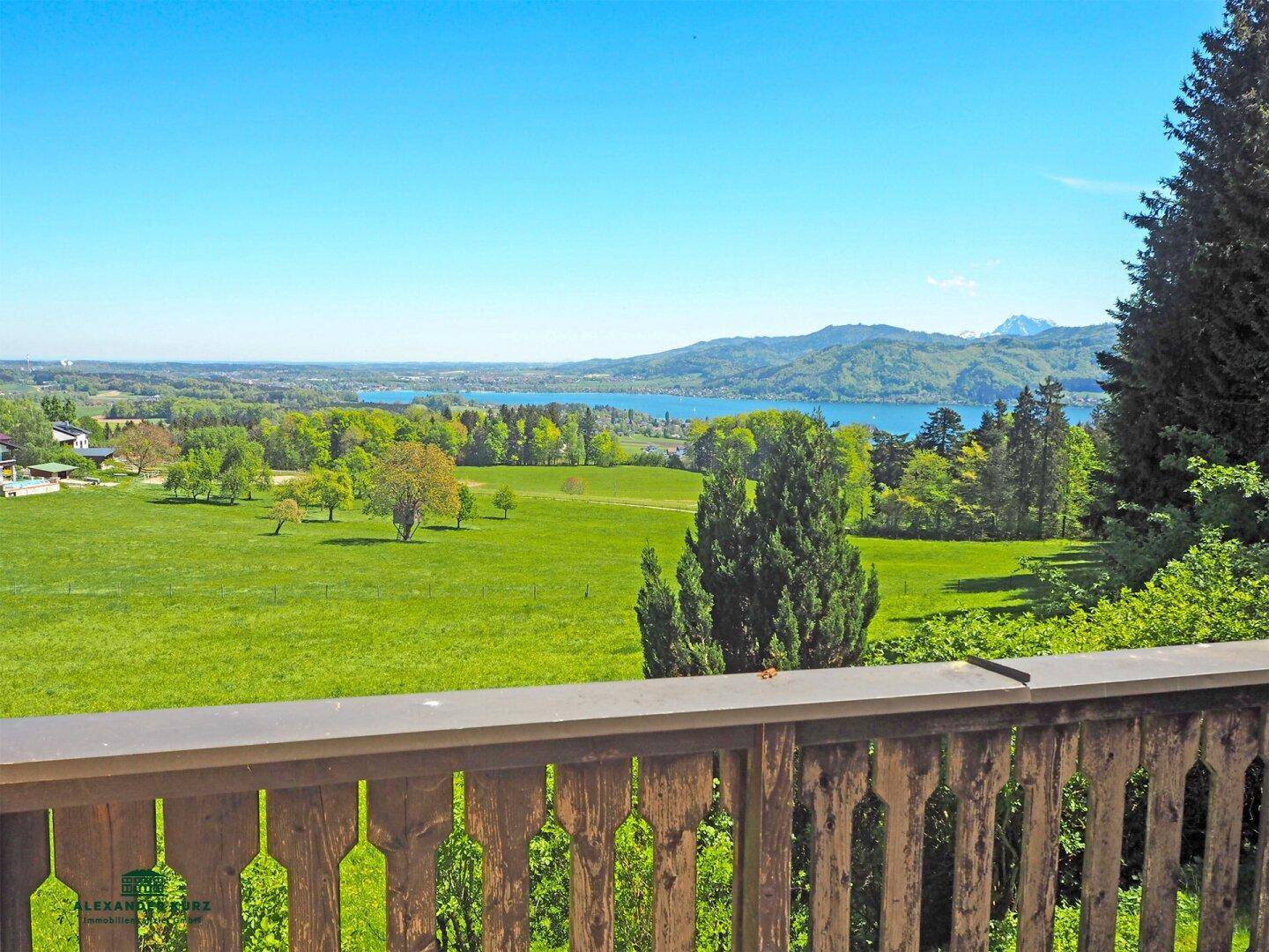 Panoramablick von einem Holzbalkon auf einen See, grüne Felder und ferne Berge unter blauem Himmel.