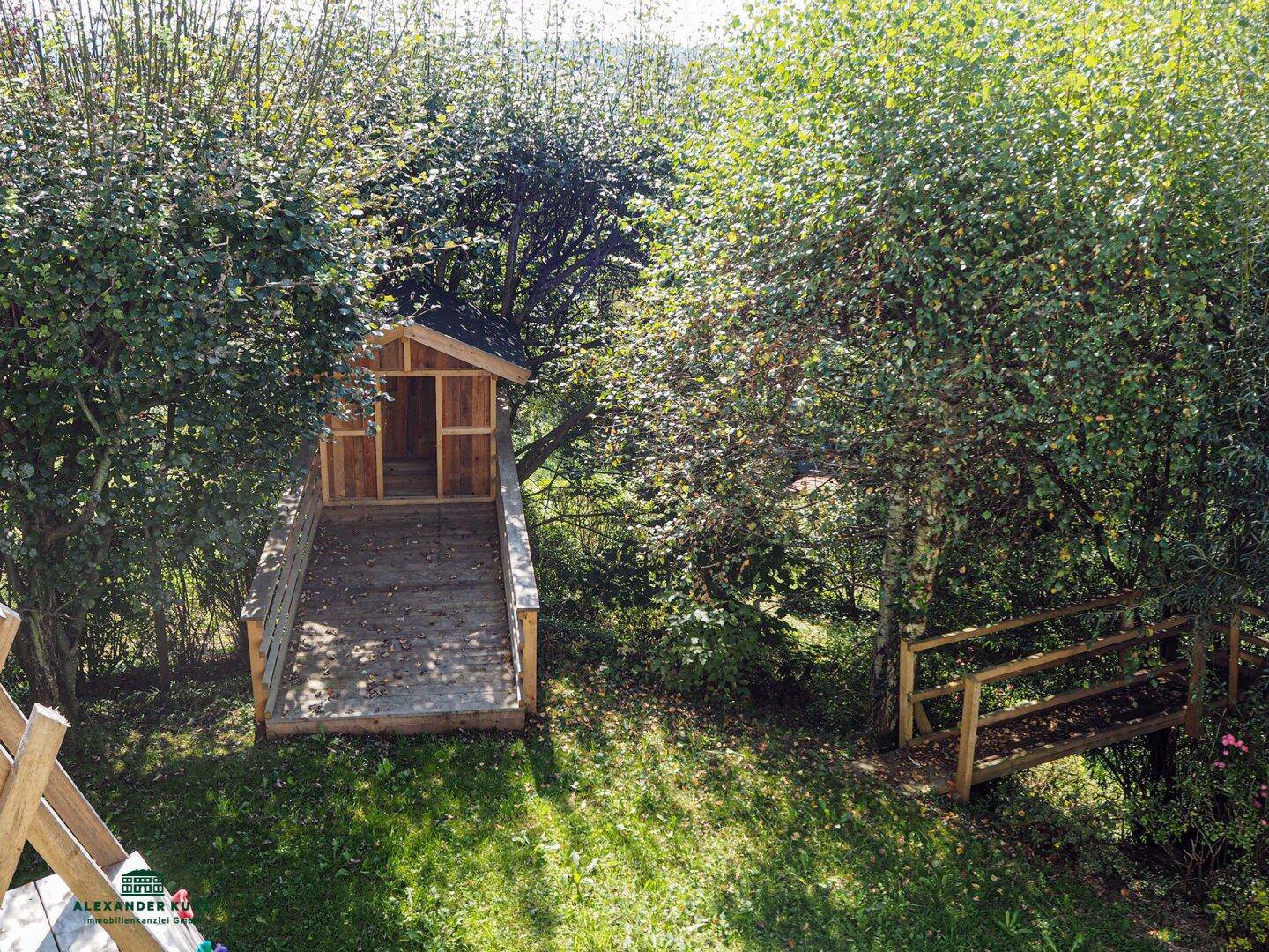 Spielhaus aus Holz mit Rampe und einer Holzbank, umgeben von Bäumen und Grünflächen im Garten.