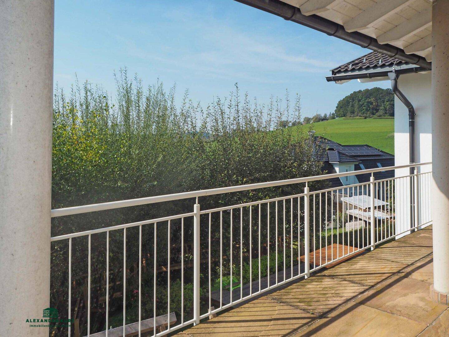 Geräumiger Balkon mit Steinboden und weißem Geländer, bietet einen weiten Blick ins Grüne.