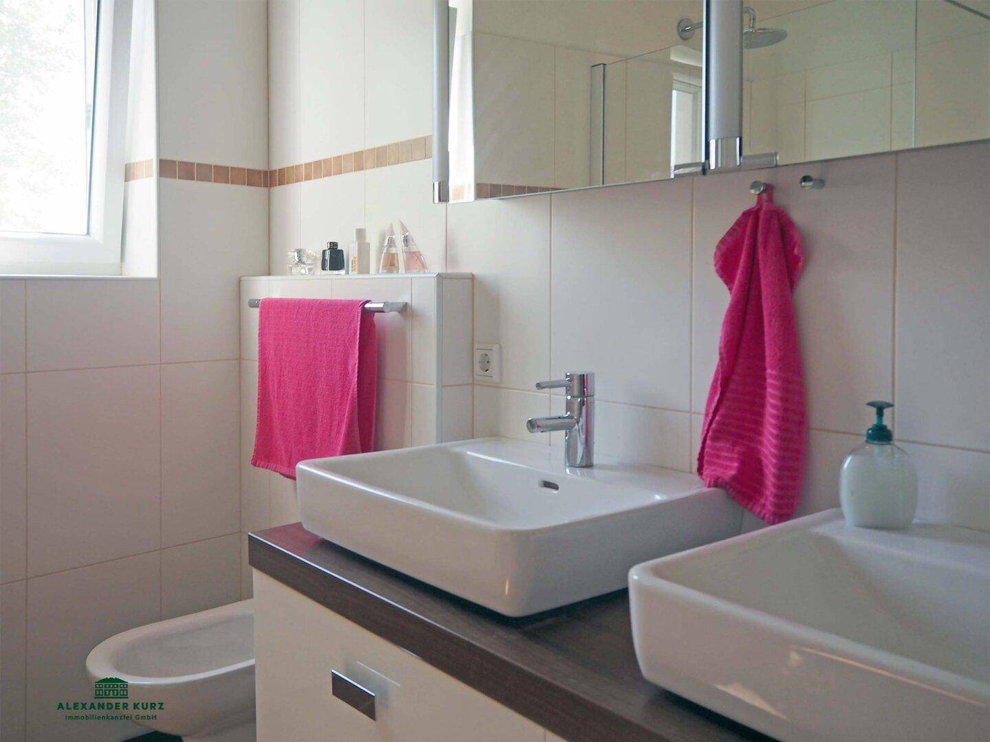 Helles Badezimmer mit zwei modernen Waschbecken, weißen Fliesen und einem Fenster für Tageslicht.