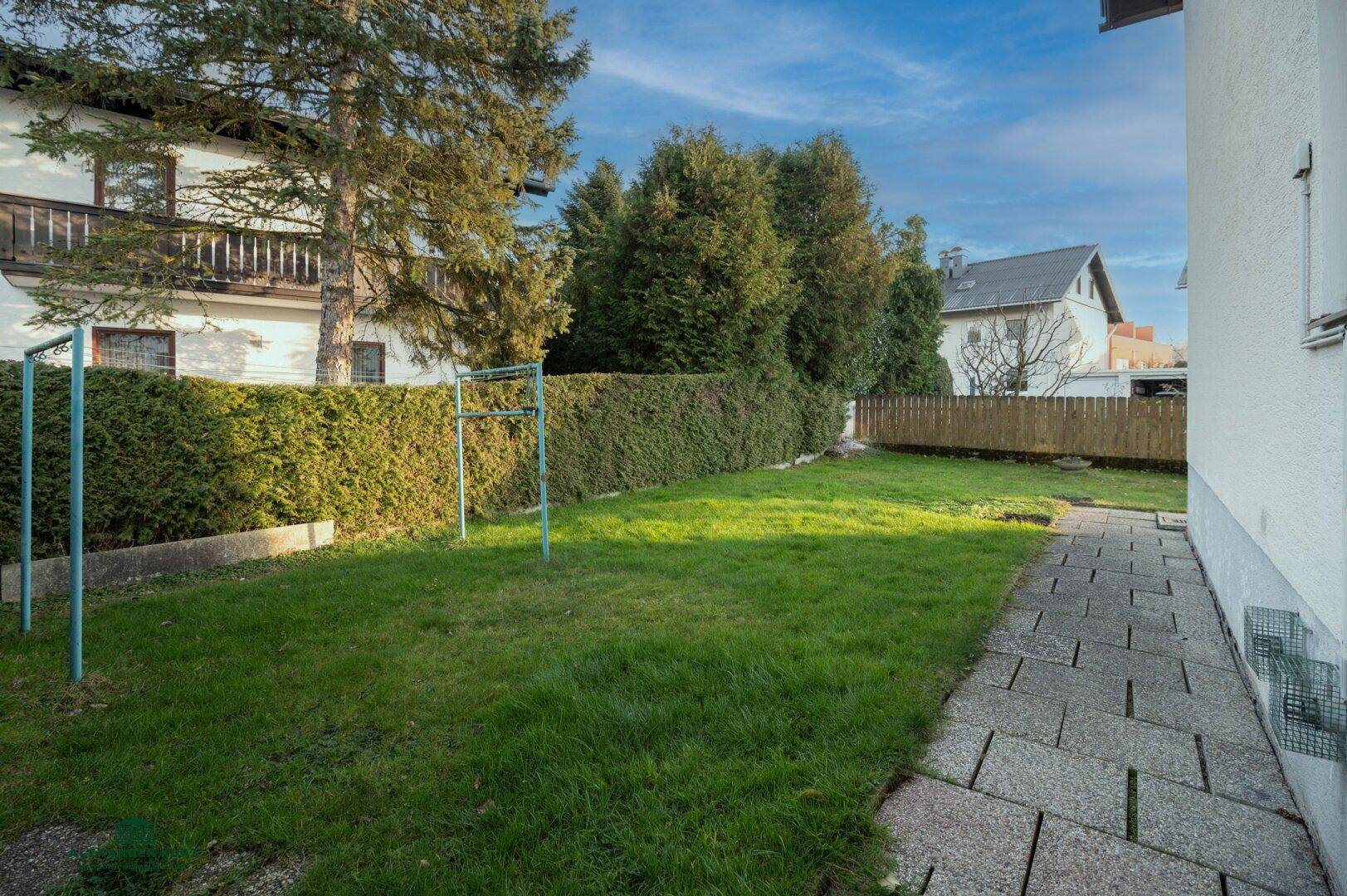 Gepflegter Gartenbereich mit Rasenfläche, hoher Hecke und Steinplattenweg.