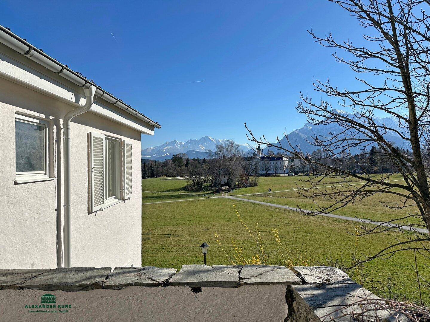 Seitenansicht des Hauses mit weitem Blick über grüne Felder und schneebedeckte Berge unter blauem Himmel.