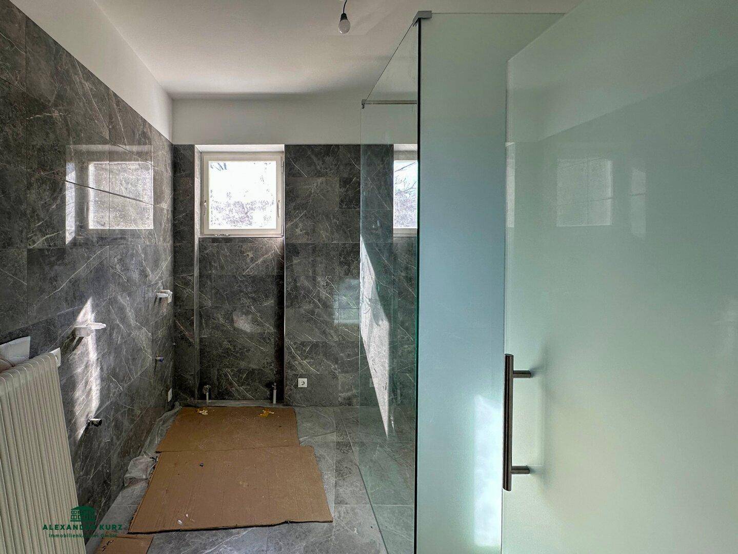 Modernes Badezimmer im Bau mit dunklen Marmorfliesen, einer großen Glasdusche und Fenster für Tageslicht.