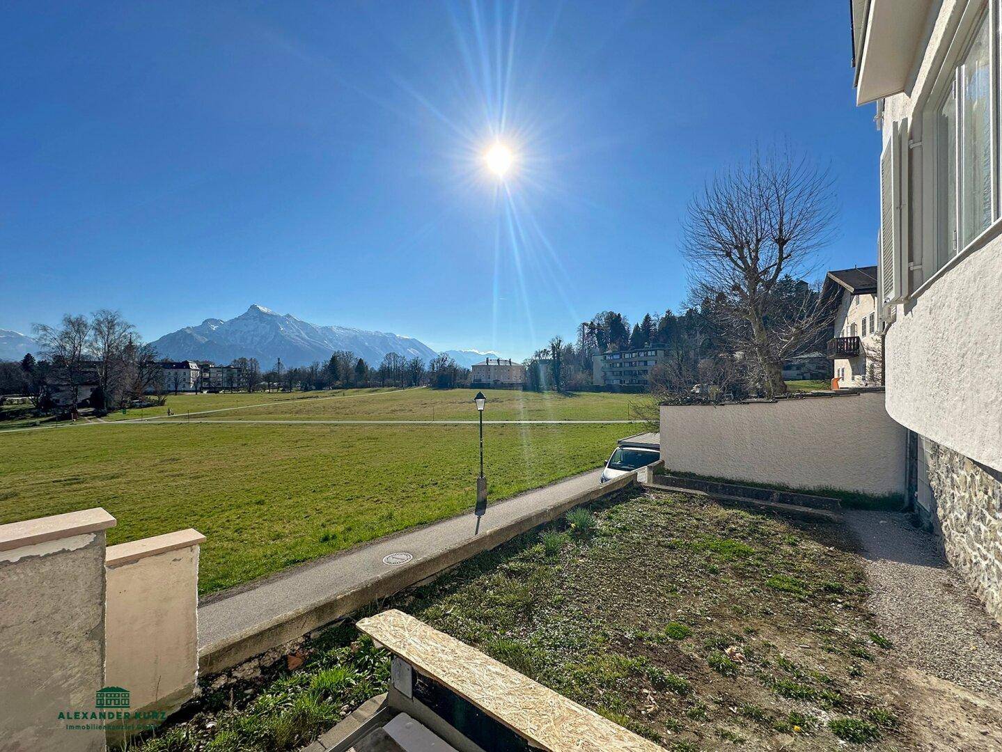 Sonniger Ausblick von der Immobilie über grüne Wiesen mit Bergen im Hintergrund und klarem Himmel.