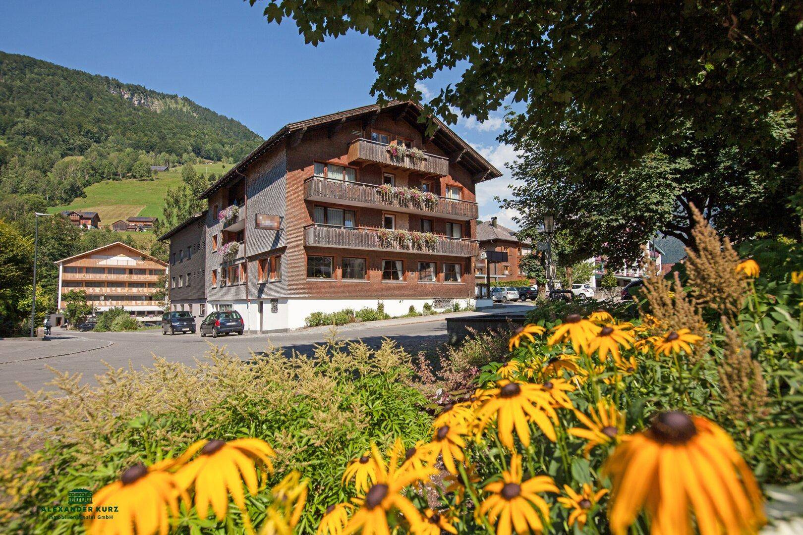 Traditionelles Gebäude im alpinen Stil, umgeben von blühenden Blumen und grünen Bergen im Sommer.