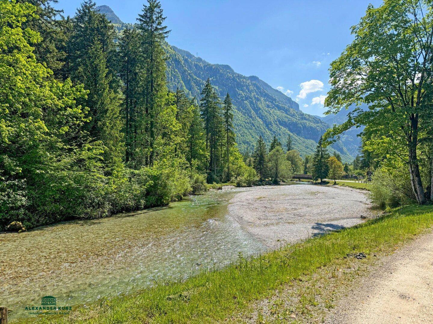 Klarer Flusslauf in einer bergigen Landschaft, umgeben von üppigen grünen Bäumen unter blauem Himmel.