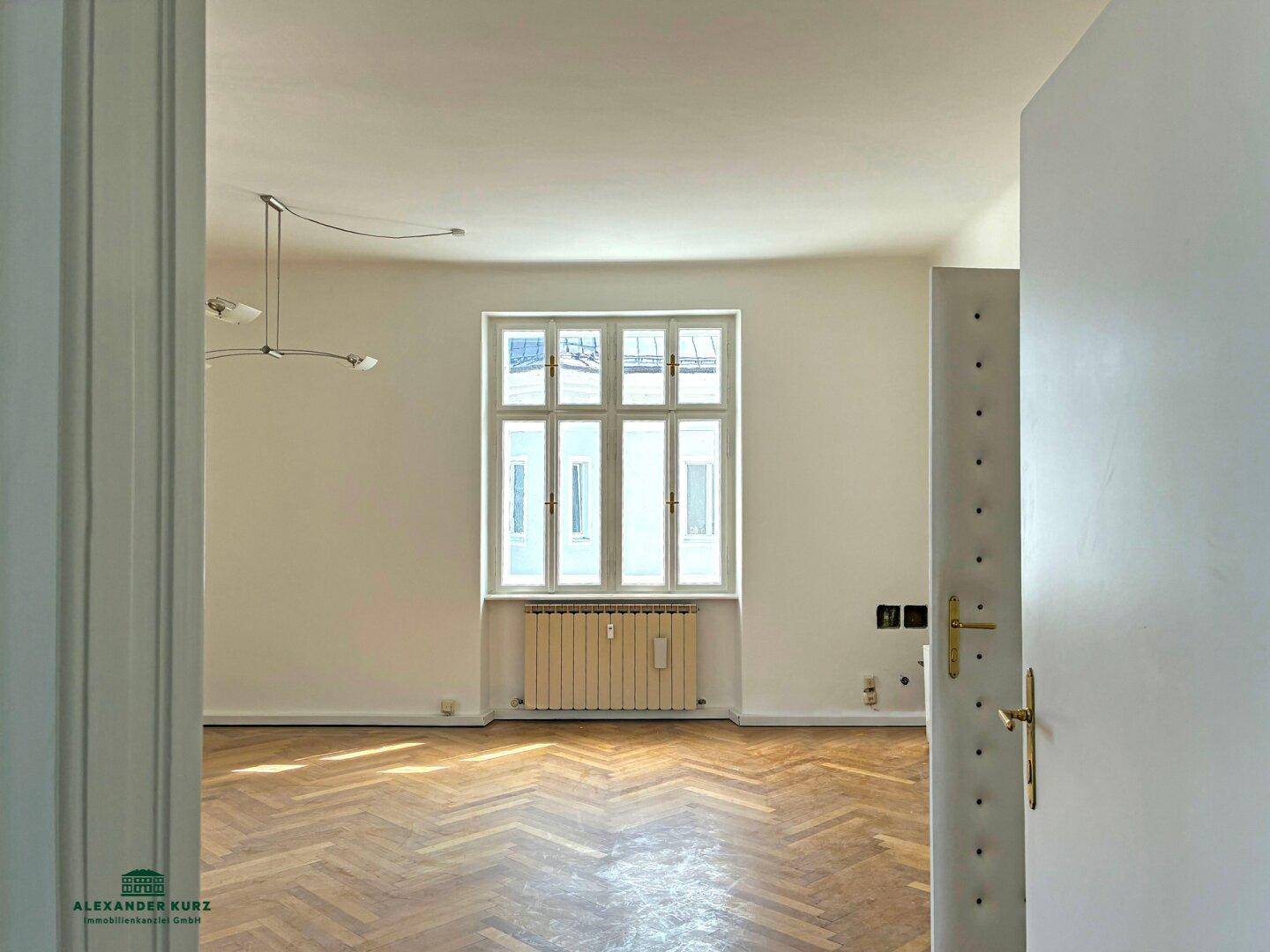 Blick durch eine offene Tür in einen hellen Raum mit Fischgrätparkett und einem großen Fenster.