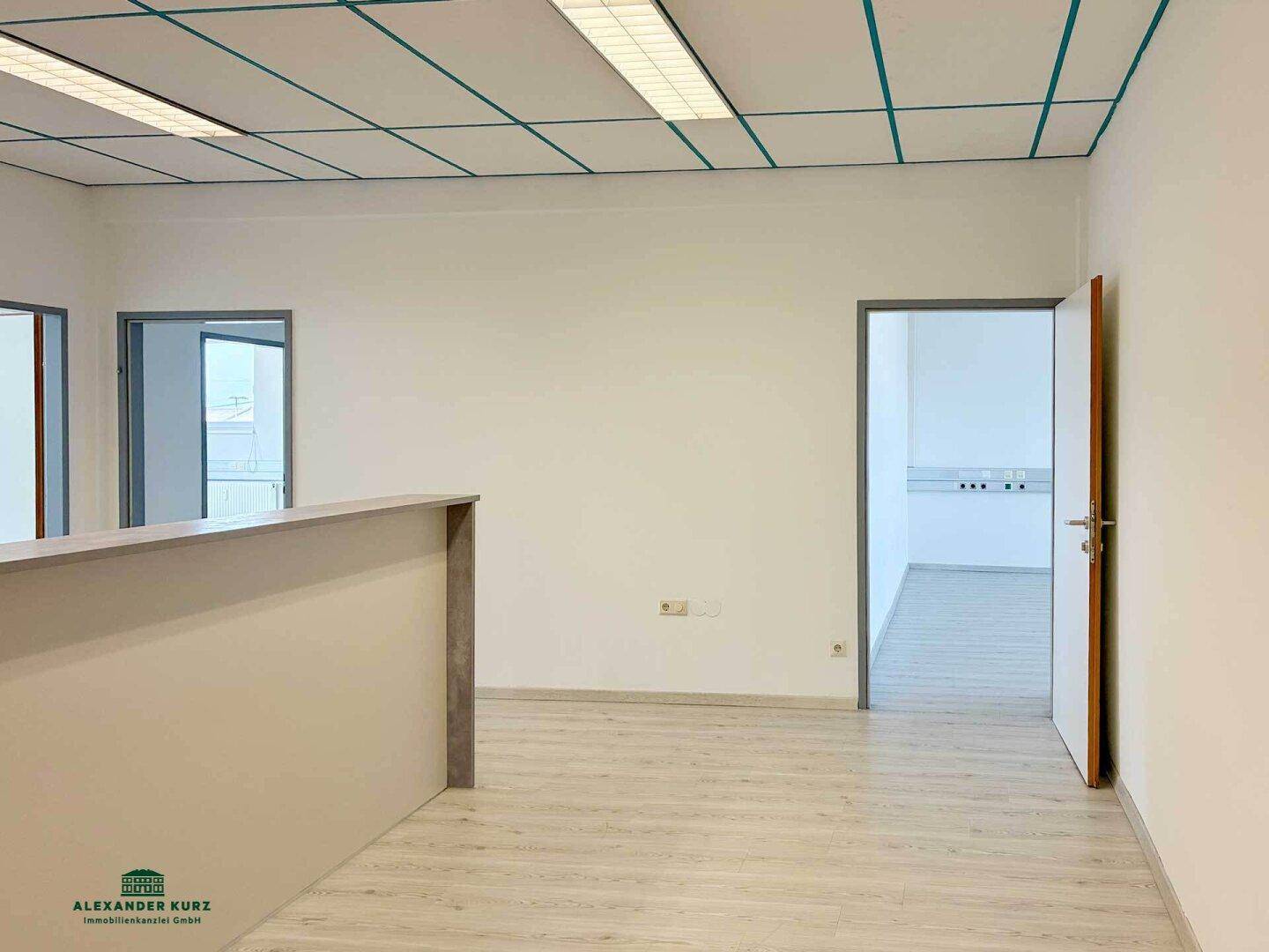 Leerer Büroraum mit Laminatboden, weißen Wänden und einer Decke mit Gitterstruktur.