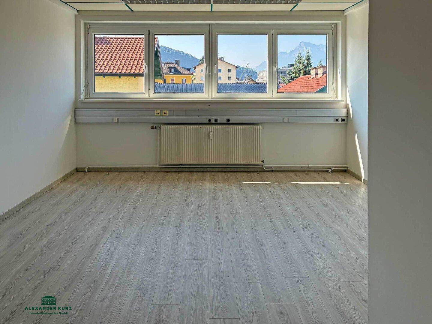 Heller Büroraum mit Laminatboden, großen Fenstern und Blick auf Berge und Dächer.