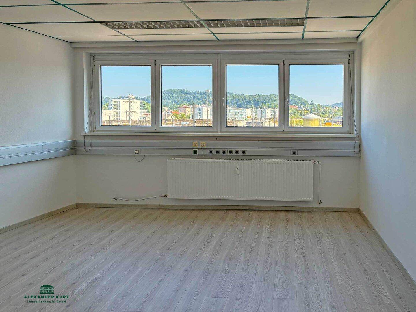 Heller Büroraum mit großen Fenstern, Laminatboden und weitem Blick ins Grüne.