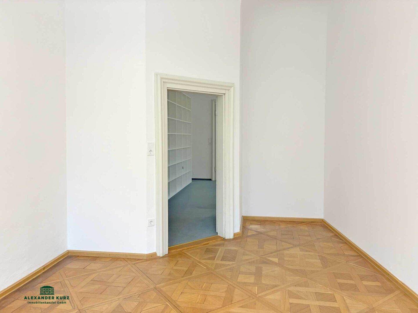 Leerer Raum mit Fischgrätparkettboden und einem Durchgang zu einem weiteren Zimmer mit Regalen.