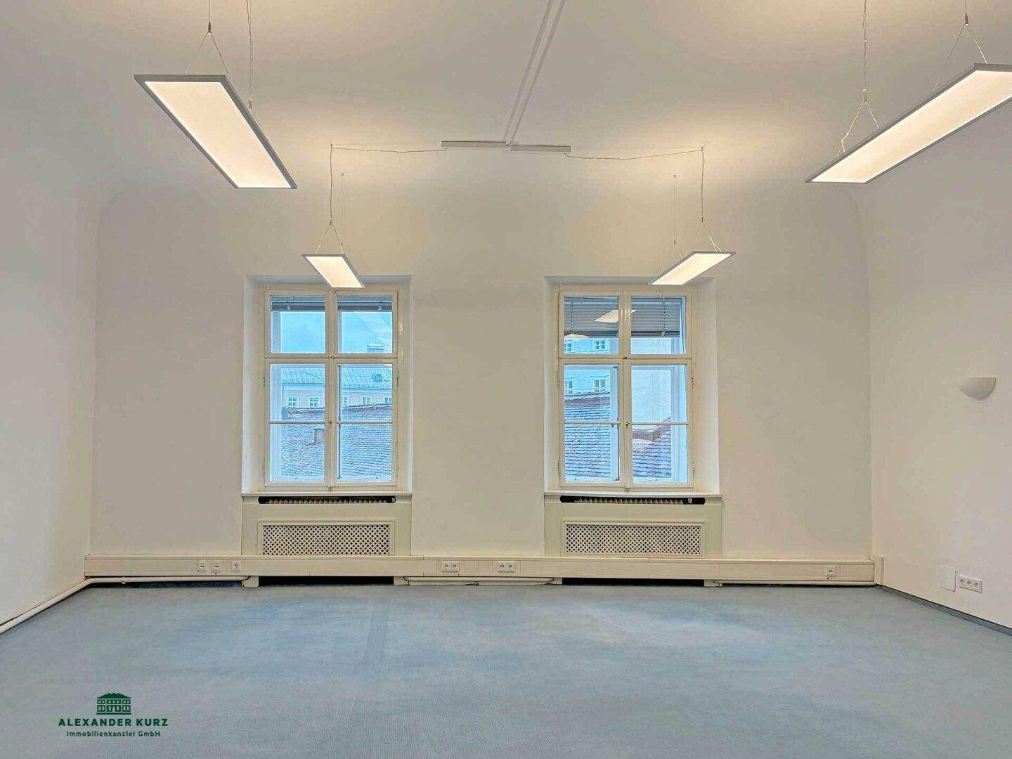 Heller Raum mit zwei großen Fenstern, modernen Deckenleuchten und blauem Teppichboden, ideal als Büro.