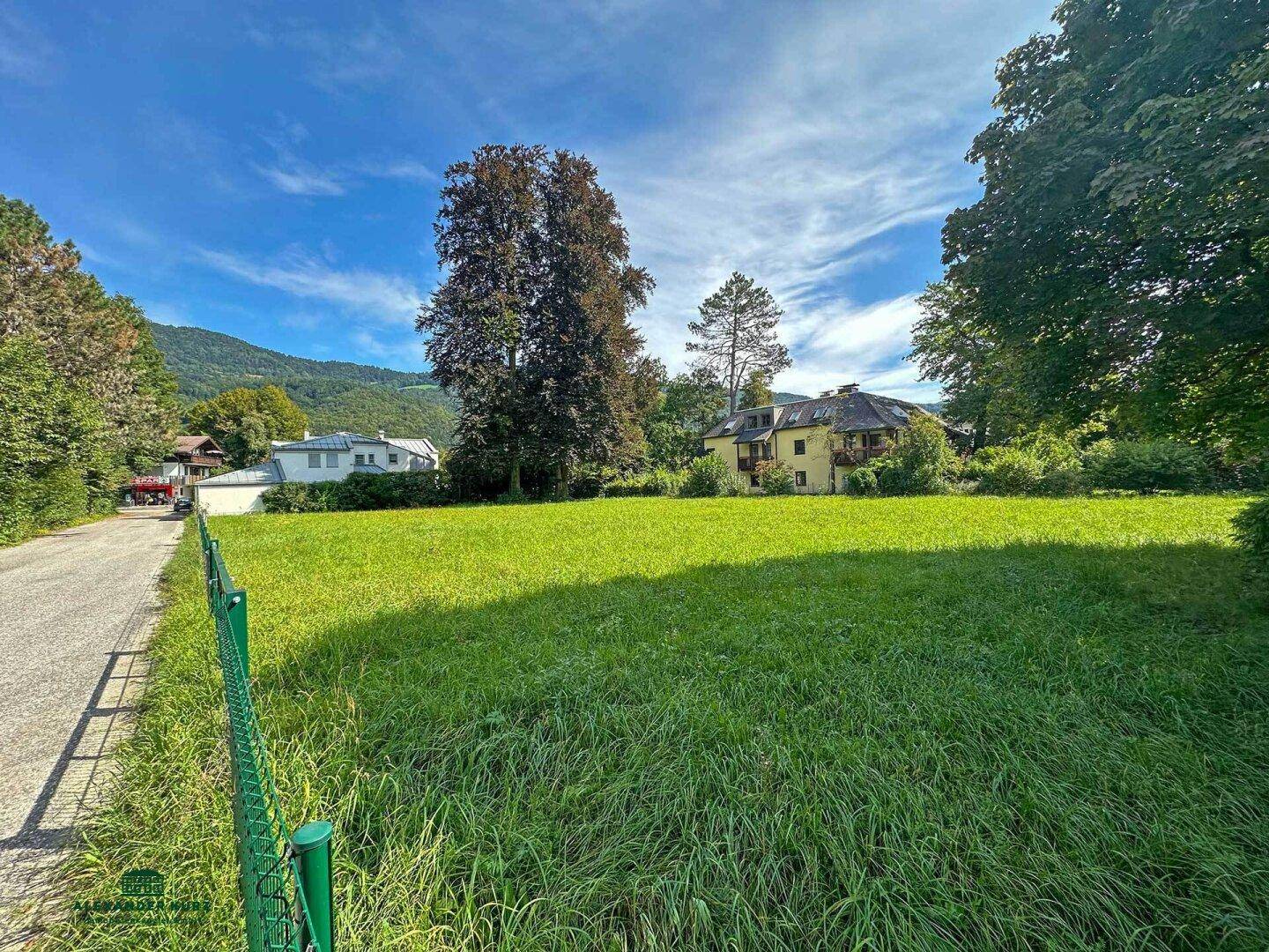 Große Wiesenfläche mit Blick auf umliegende Häuser und bewaldete Berge im Hintergrund.