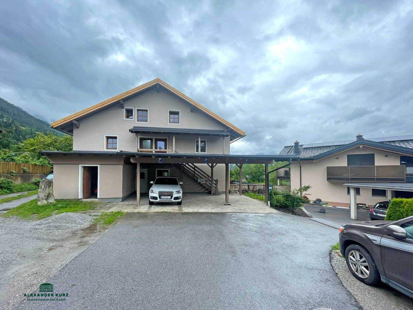 Rückansicht des Hauses mit Carport und Zufahrt, umgeben von grüner Landschaft und Bergen.