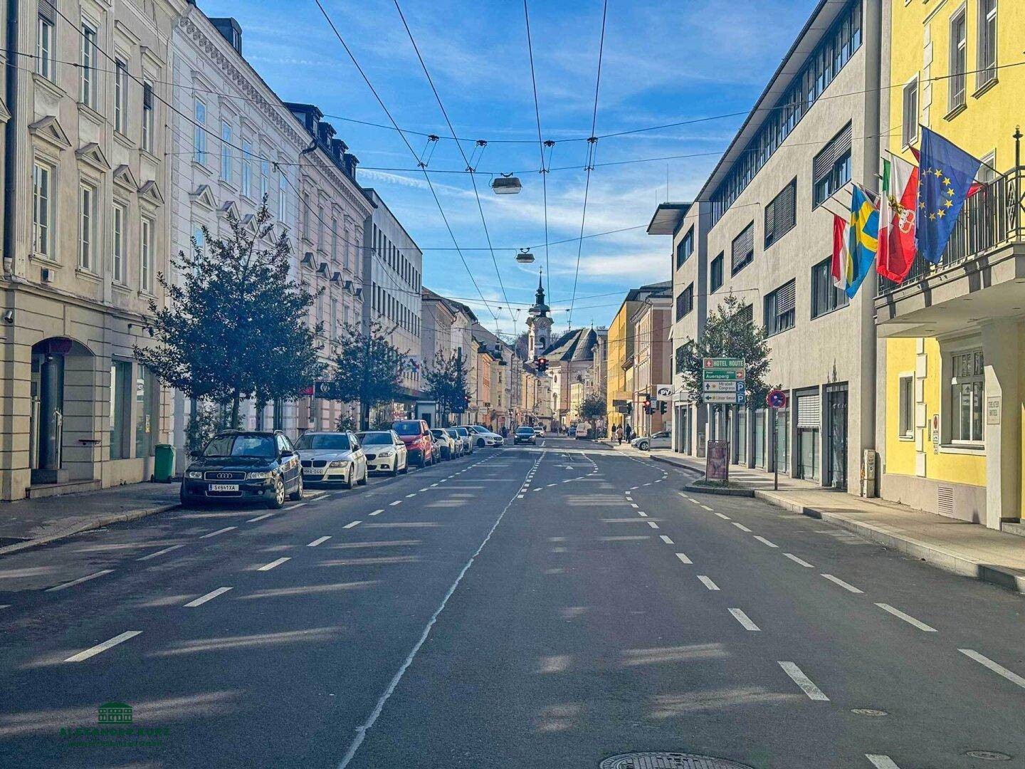 Weitläufige Stadtstraße mit parkenden Autos und Gebäuden unter klarem blauem Himmel.