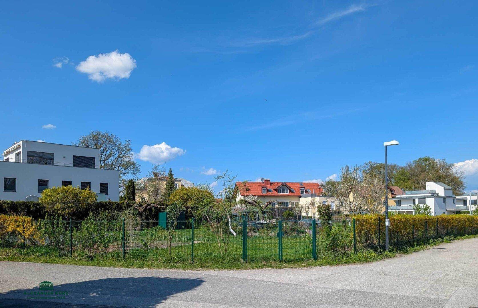 Blick auf ein unbebautes Grundstück mit umliegenden Wohnhäusern und viel Grün unter blauem Himmel.