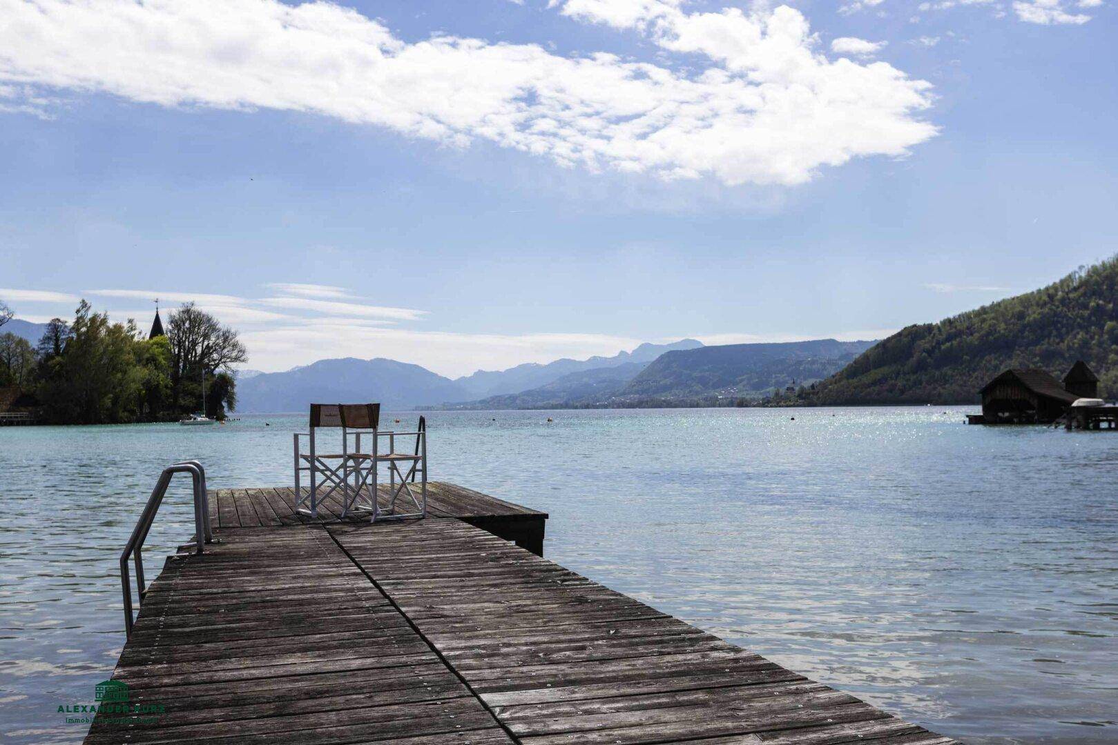 Idyllischer Seeblick mit Holzsteg und majestätischer Bergkulisse unter blauem Himmel.