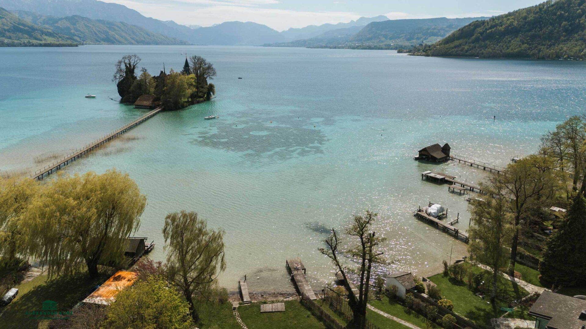 Panoramablick auf den See mit kleiner Insel, Stegen und umliegender grüner Landschaft.